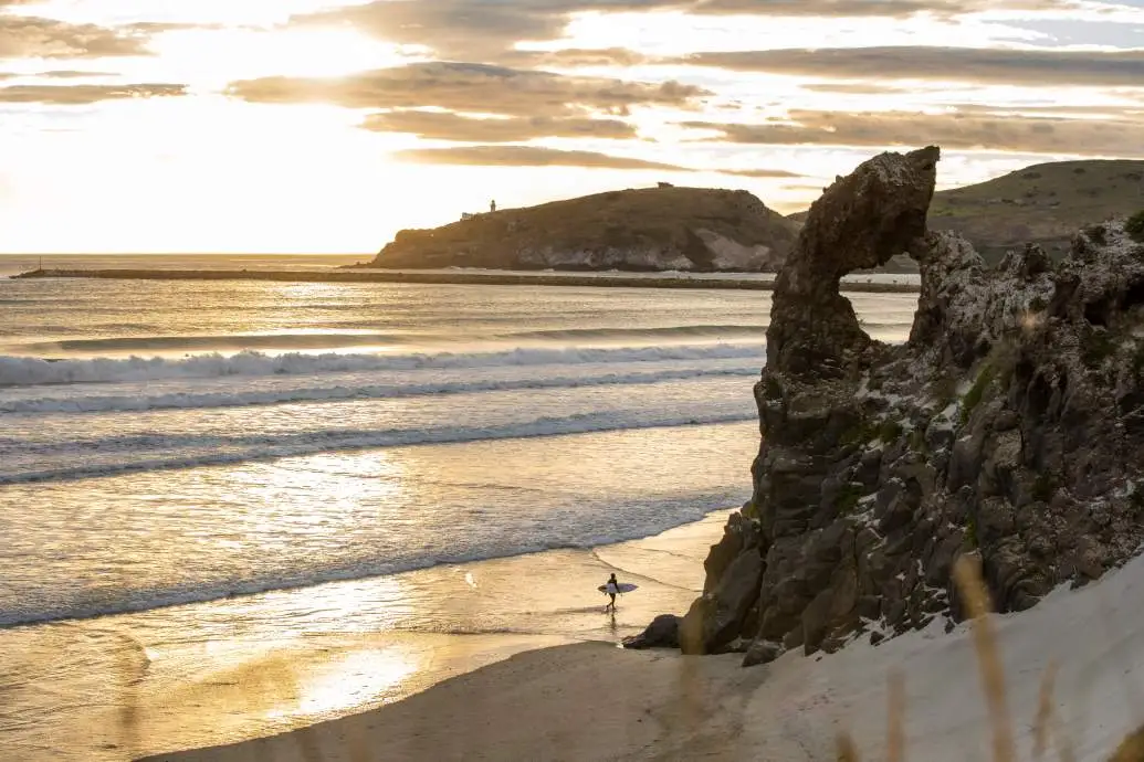 A surfer walks along Aramoana Beach beneath Bear Rock at sunset.