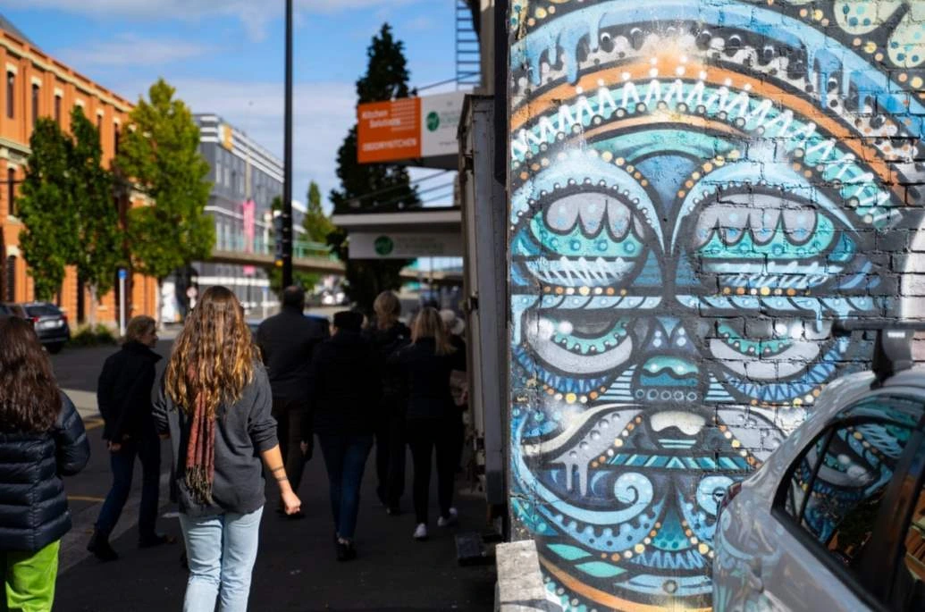 A group of people walk down a street with blue-green street art on a brick wall to their right.