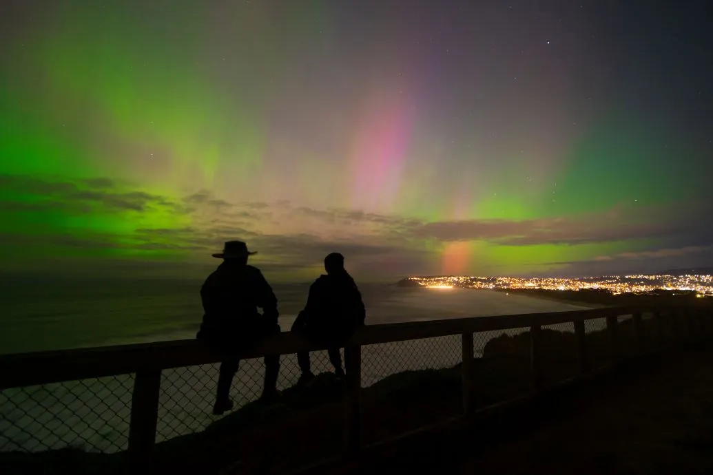 Two men watch the Southern Lights above the city at ngiht.