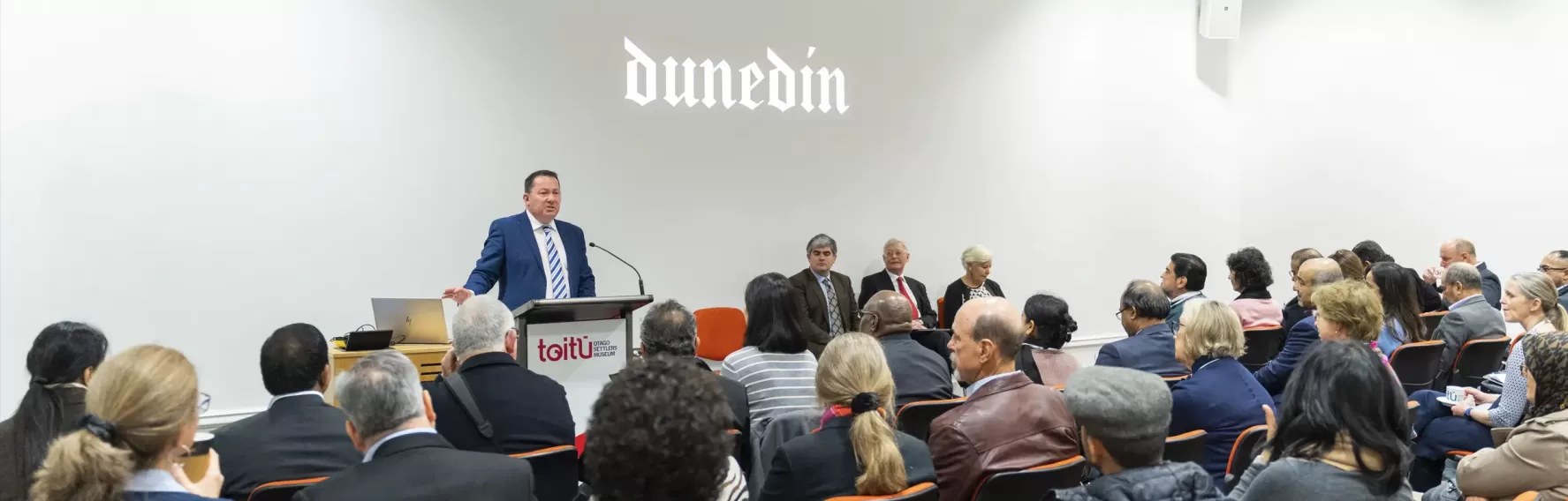 A crowd of people are seated facing towards a man speaking at a podium at the front of the room. The word Dunedin in a gothic font is projected onto the white wall beside him 