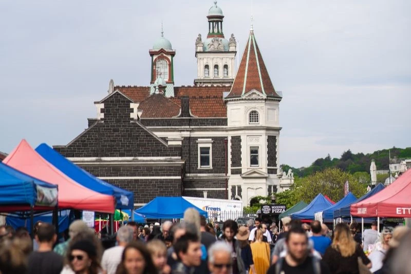 A bustling crowd of people at the Otago Farmers Market with the Dunedin Railway Station in the background.