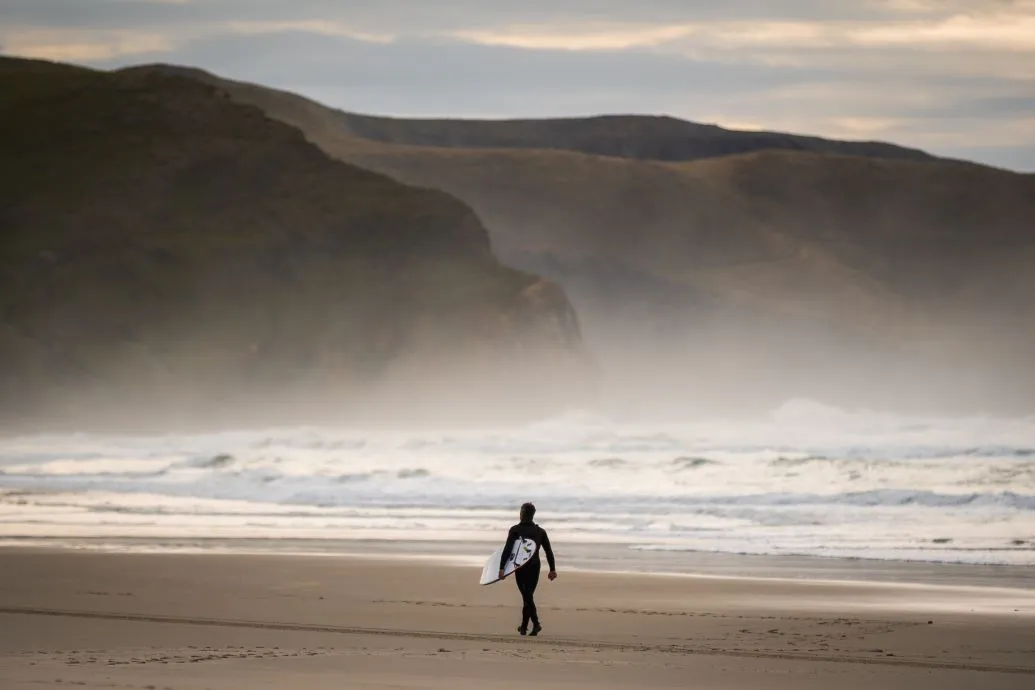 A surfer walks along the mist Allans Beach.