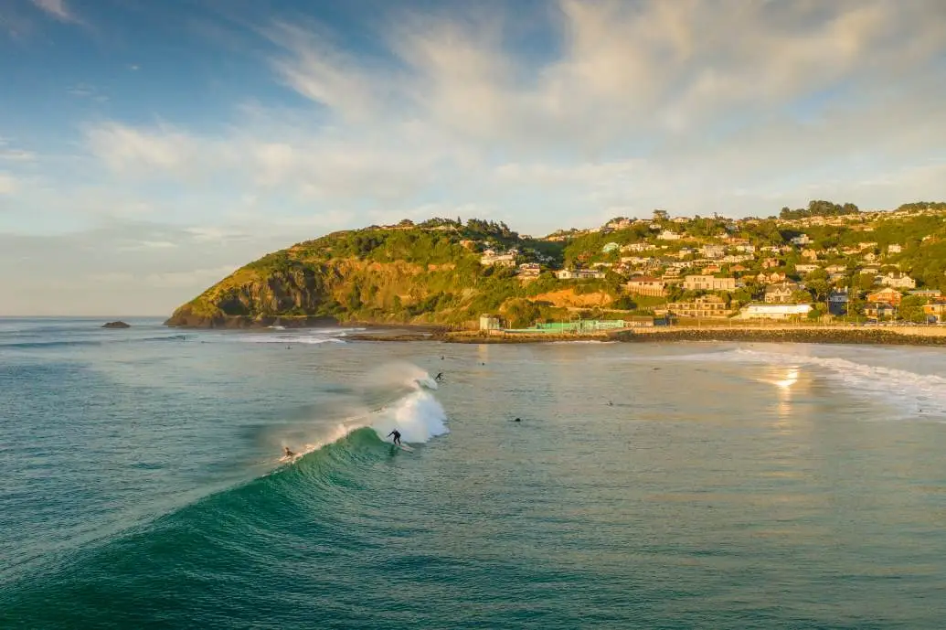 St Clair bathed in morning sunlight with a surfer catching a wave.