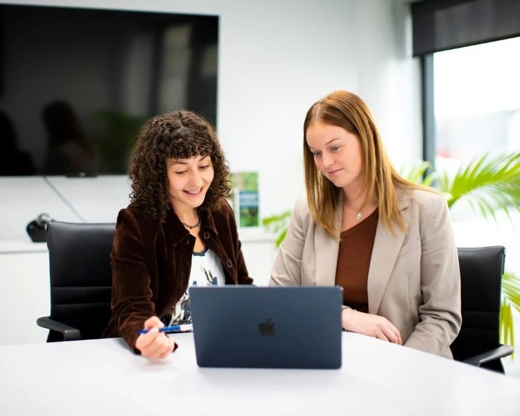 Two women discuss their work in front of a laptop.