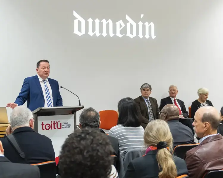 A crowd of people are seated facing towards a man speaking at a podium at the front of the room. The word Dunedin in a gothic font is projected onto the white wall beside him 