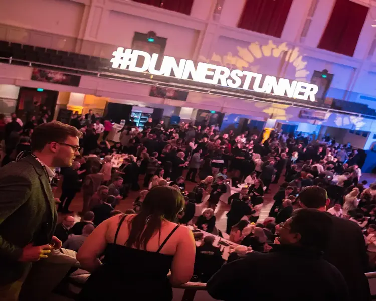 A large crowd enjoy themselves in front of a stage with sound and lighting equipment in full swing. A light display along the side of the venue says DUNNERSTUNNER 