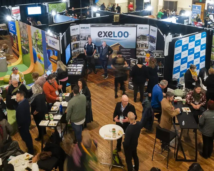 A large number of people congregate in groups of two to four. They are moving around colourful trade displays and catering tables. 