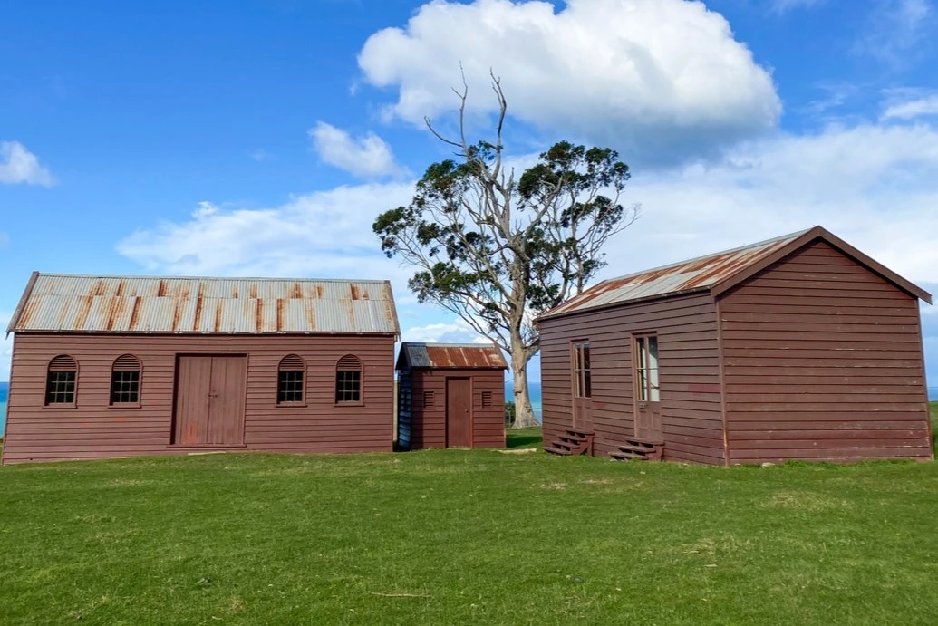Matanaka Farm Three red-brown heritage structures, the original settler farm buildings stand on a green lawn.